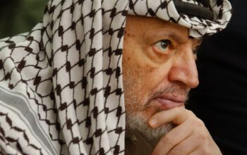 Potret jarak dekat Yasser Arafat mengenakan keffiyeh ikoniknya, menatap ke kejauhan dengan ekspresi berpikir yang mendalam, menyoroti kisah hidupnya sebagai seorang pemimpin.