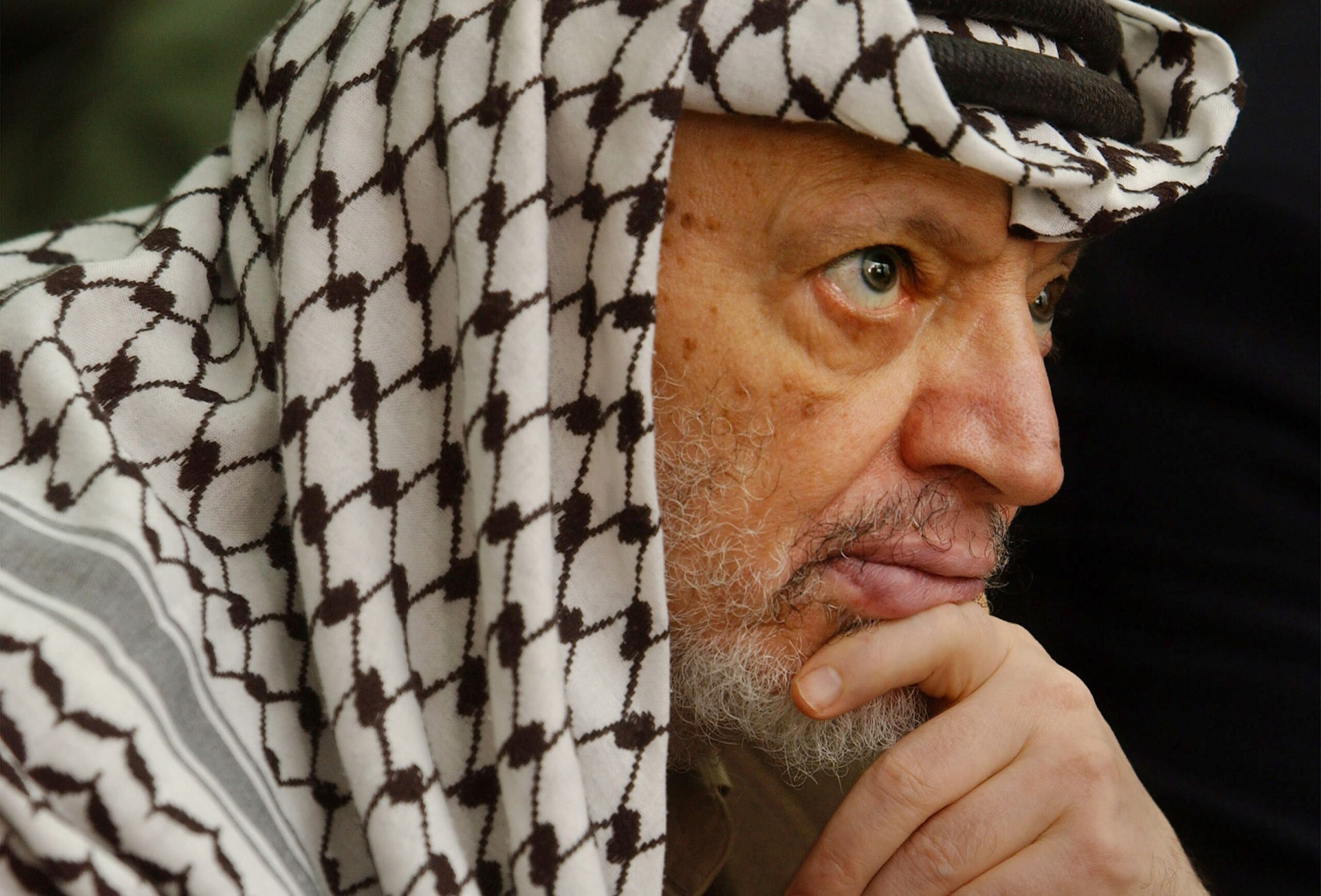 Potret jarak dekat Yasser Arafat mengenakan keffiyeh ikoniknya, menatap ke kejauhan dengan ekspresi berpikir yang mendalam, menyoroti kisah hidupnya sebagai seorang pemimpin.