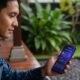 Pria Indonesia berusia 30-an dengan kemeja batik biru motif etnis duduk di teras kayu rumah tropis, memegang smartphone Samsung berwarna hitam dengan layar menampilkan aplikasi cuaca AI "Weekly Weather History" (grafik suhu, curah hujan, dan ikon matahari). Latar belakang: tanaman hijau, rumput, dan elemen arsitektur tradisional Indonesia.